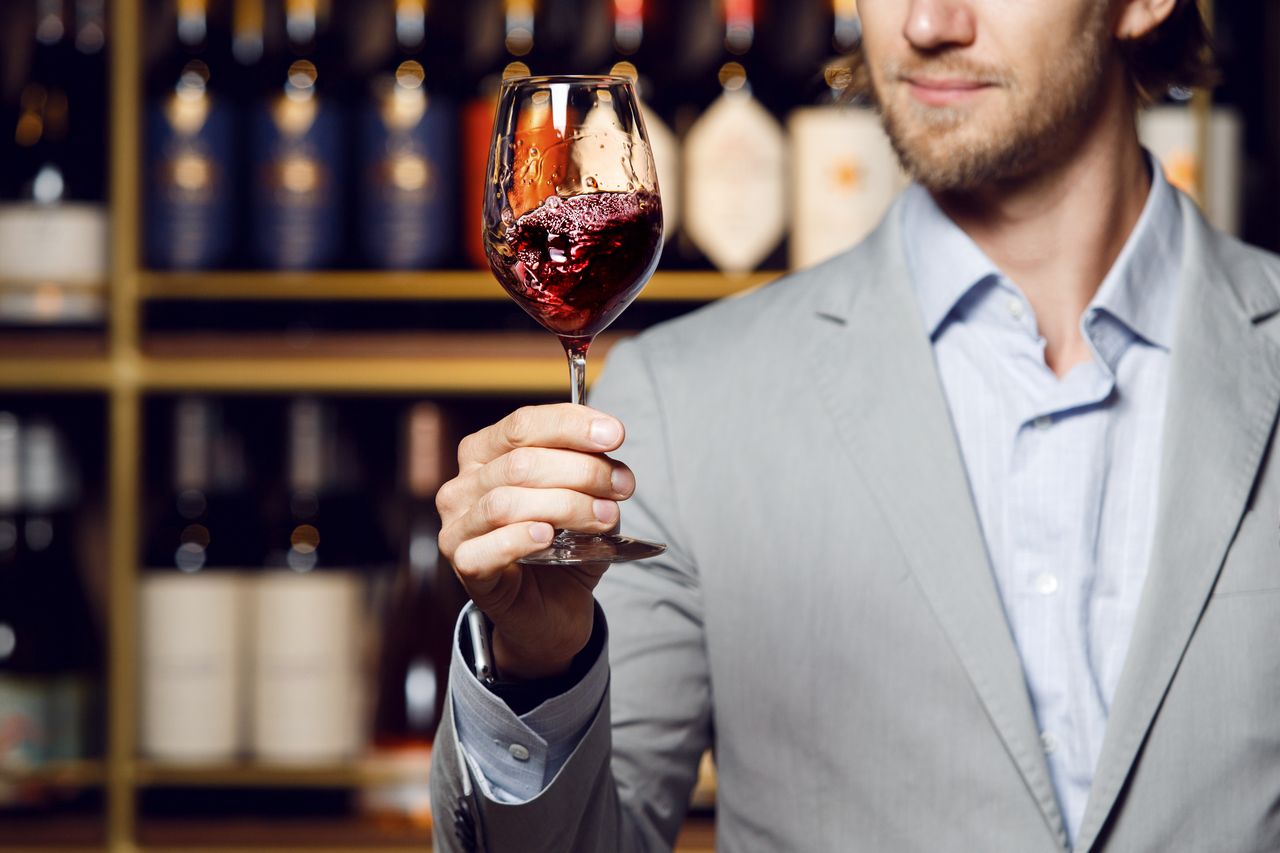 Homme d'une cinquantaine d'années aux tempes grises, chemise en lin, verre de vin rouge à la main dans une cave voûtée chaleureusement éclairée